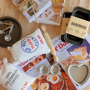 May include: A collection of miniature baking supplies, including a bag of flour, a bag of coffee, a rolling pin, baking pans, measuring spoons, and a heart-shaped mold. The scene is set on a vintage-style magazine page with the word "BAKEWARE" on a label.