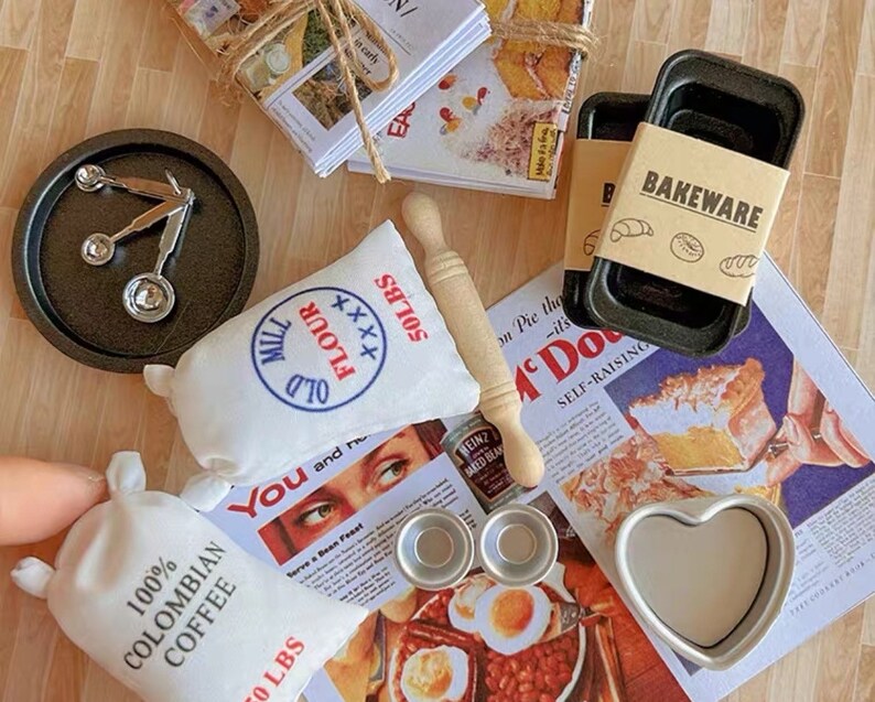 May include: A collection of miniature baking supplies, including a bag of flour, a bag of coffee, a rolling pin, baking pans, measuring spoons, and a heart-shaped mold. The scene is set on a vintage-style magazine page with the word "BAKEWARE" on a label.