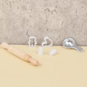 May include: A collection of baking tools including a wooden rolling pin, white plastic cookie cutters in bear and heart shapes, two white piping tips, and a silver egg separator. The tools are arranged on a light-colored surface.