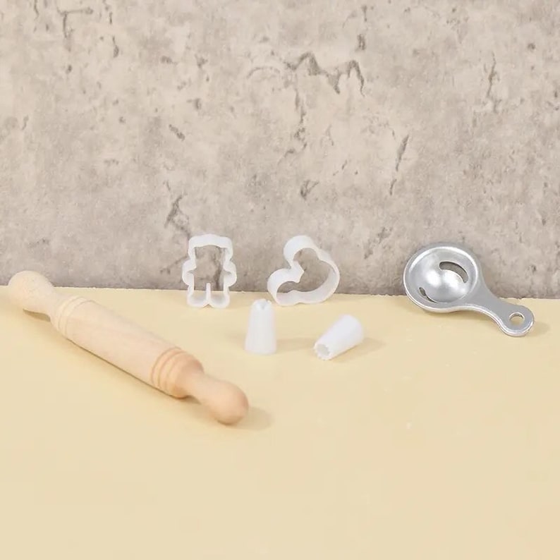 May include: A collection of baking tools including a wooden rolling pin, white plastic cookie cutters in bear and heart shapes, two white piping tips, and a silver egg separator. The tools are arranged on a light-colored surface.