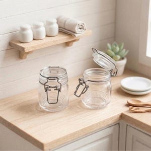 Puede incluir: Dos frascos de vidrio transparente con cierres metálicos, uno abierto y otro cerrado, sobre una superficie de madera clara. Una pequeña estantería de madera encima contiene saleros de cerámica blanca y una toalla beige enrollada. Una escena de cocina en miniatura.