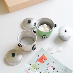 May include: A set of miniature rice cookers with lids and a small measuring cup and spatula. The cookers are white, green, and silver. One cooker has a white plate with three small white buns inside.