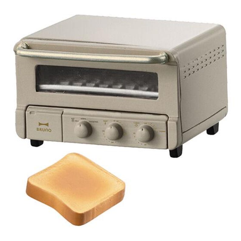 May include: A beige miniature toaster oven with a glass door, a silver handle, and three control knobs on the front panel featuring the brand name BRUNO. A single piece of light brown square toast is positioned in front of the appliance.