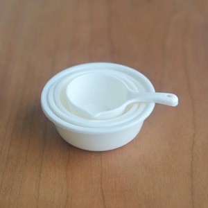 Peut inclure: Un ensemble de quatre tasses à mesurer blanc cassé et une petite cuillère à mesurer. Les tasses sont emboîtées les unes dans les autres, avec la cuillère posée dessus. Les articles sont en matériau lisse et solide.