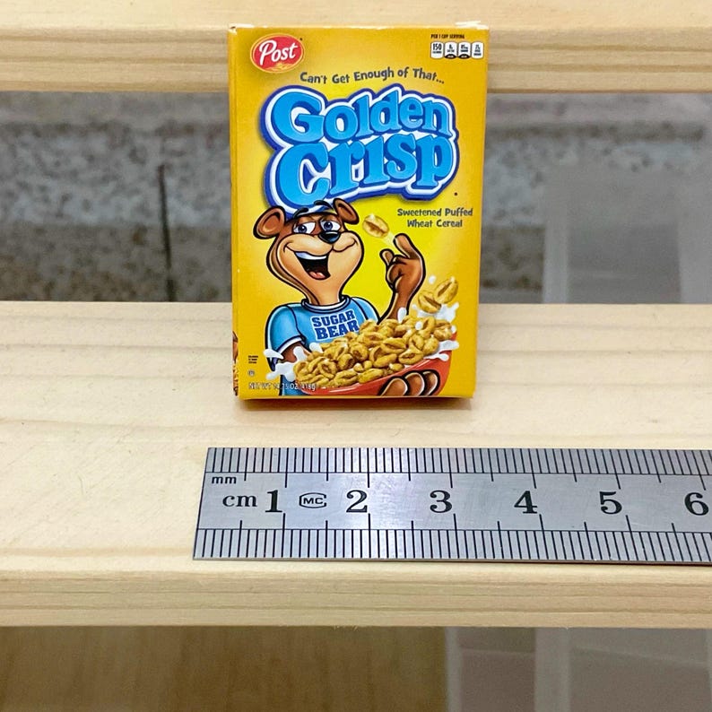 May include: A miniature box of Golden Crisp cereal. The box is yellow with blue and white text, and features a cartoon bear mascot. The cereal box is next to a ruler, with measurements in centimeters.