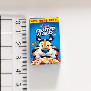 May include: Miniature Kellogg's Frosted Flakes cereal box. The box is blue with the Frosted Flakes logo and Tony the Tiger. The top of the box reads "42% MORE FREE". The cereal box is next to a ruler.