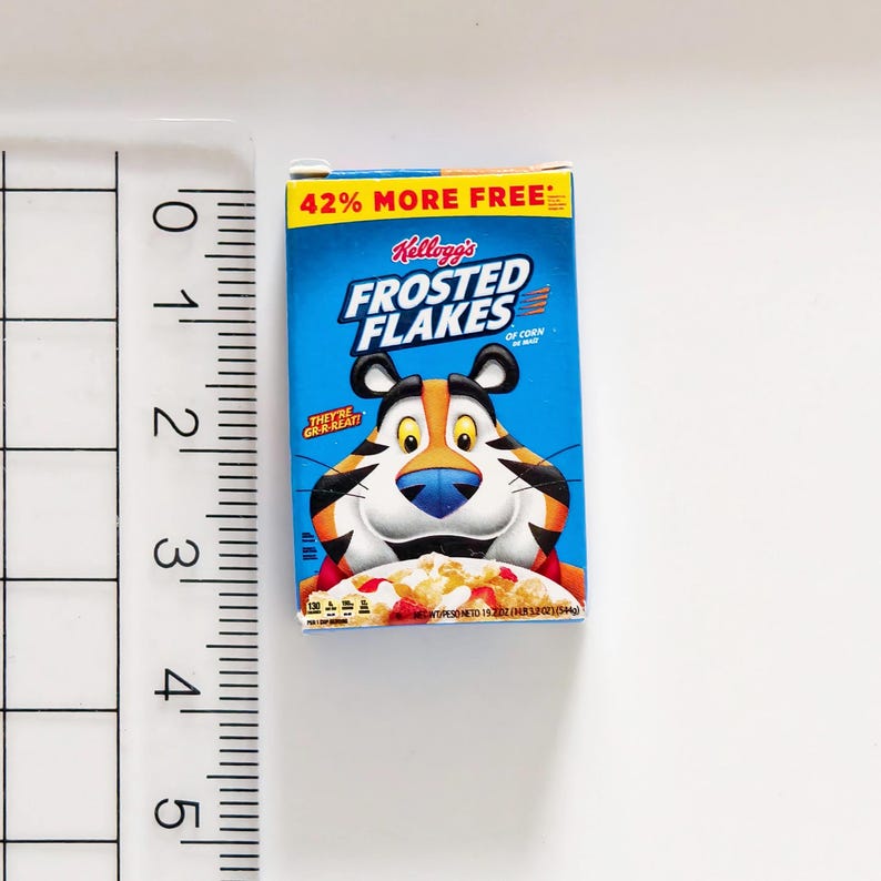 May include: Miniature Kellogg's Frosted Flakes cereal box. The box is blue with the Frosted Flakes logo and Tony the Tiger. The top of the box reads "42% MORE FREE". The cereal box is next to a ruler.