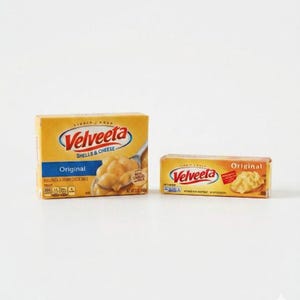Könnte beinhalten: Zwei Schachteln Velveeta Shells & Cheese. Eine Schachtel ist rechteckig, die andere ziegelartig. Beide sind gelb mit dem Velveeta-Logo und den Wörtern "Original" und "Shells & Cheese" aufgedruckt.