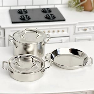 May include: A set of miniature stainless steel cookware, comprising a lidded pot, a smaller lidded pot, and a frying pan. Ideal for a doll's house or miniature kitchen. The background features a black hob and white kitchen cabinets.