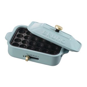 May include: A light blue electric takoyaki maker with a rectangular shape and a matching lid. The lid features the embossed text BRUNO and a gold-colored knob. The base has a black non-stick plate with twenty-four circular molds and a gold slider.