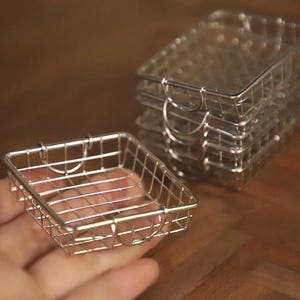 May include: A set of small, square, wire mesh baskets. One basket is held in a hand, while a stack of similar baskets sits in the background. The baskets have a shiny, metallic finish and a simple, minimalist design.