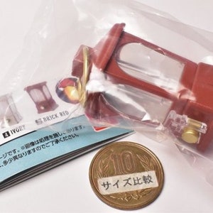 May include: A red and clear plastic miniature lantern with a gold-colored handle. The lantern is sitting on a white surface with a piece of paper that says "IVORY" and "BRICK RED". There is also a Japanese 100 yen coin with the text "サイズ比較" on it.