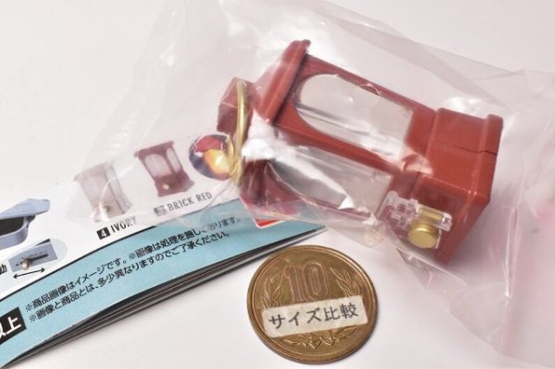 May include: A red and clear plastic miniature lantern with a gold-colored handle. The lantern is sitting on a white surface with a piece of paper that says "IVORY" and "BRICK RED". There is also a Japanese 100 yen coin with the text "サイズ比較" on it.