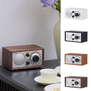 May include: A collection of small, retro-style radios in various colors, including wood grain, white, black, and brown. Each radio features a silver speaker grill, control knobs, and a tuning dial. The radios are displayed on a dark surface.