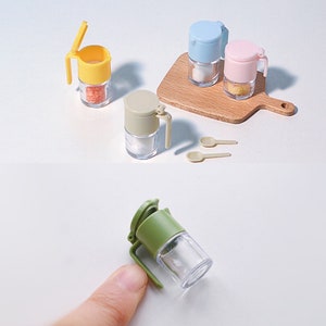 Può includere: Set di cinque barattoli per spezie in miniatura con coperchi e manici. I barattoli sono in vetro trasparente con coperchi e manici in plastica colorata. I coperchi sono blu, rosa, giallo, verde e beige. I barattoli sono su un tagliere di legno con due piccoli cucchiai.