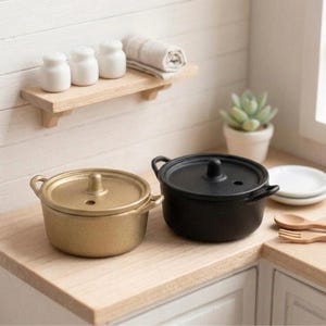 May include: Miniature gold and black cooking pots with lids. The gold pot is on the left, and the black pot is on the right. A small wooden shelf holds white salt shakers and a rolled-up towel. A small succulent plant is in the background.