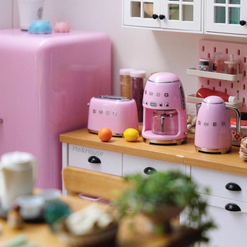 Peut inclure: Un ensemble de cuisine rose miniature comprenant un r&eacute;frig&eacute;rateur, un grille-pain, une cafeti&egrave;re et une bouilloire. Les appareils sont dispos&eacute;s sur un comptoir en bois avec un meuble blanc. Deux fruits sont sur le comptoir. Le mot "SMEG" est visible.