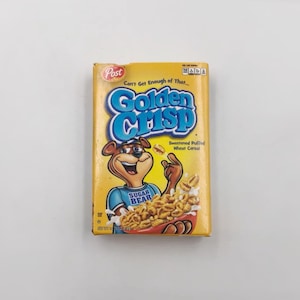 May include: A yellow box of Golden Crisp cereal. The box features a cartoon bear wearing a blue shirt that says "Sugar Bear." The cereal name is in large blue letters. The box also says "Sweetened Puffed Wheat Cereal."