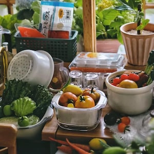 May include: A collection of kitchen items including a white colander filled with green leafy vegetables, a clear glass square container holding tomatoes and a lemon, a white bowl with tomatoes, and a plastic egg carton on a wooden surface.