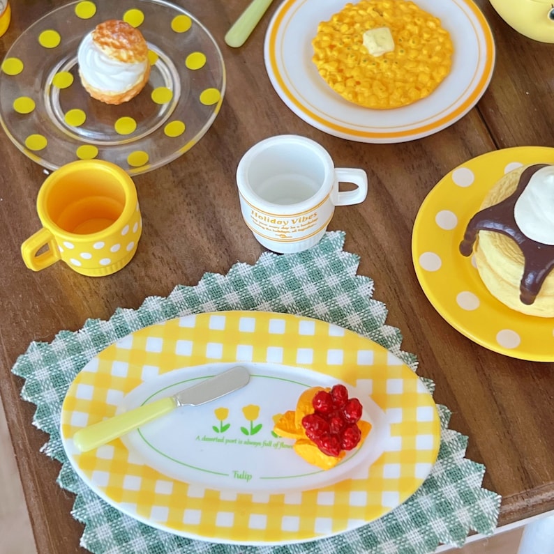 Op de afbeelding: Een geel en wit geruit bord met een klein mes en een portie fruit. Het fruit is een mix van rode frambozen en sinaasappelschijfjes. Het bord ligt op een groen en wit geruit placemat. De tekst op het bord luidt "A delightful part is always full of flower - Tulip".