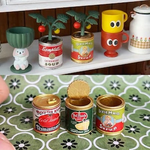 Pode incluir: Três latas em miniatura com rótulos vintage. As latas são rotuladas com "Campbell's Chicken Noodle Soup", "Dixie Maid Syrup" e "Del Monte Florida Grapefruit Juice".