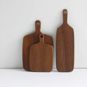 Puede incluir: Tres tablas de cortar de madera de diferentes tamaños con asas. Las tablas son todas de madera marrón oscura y tienen un patrón de veta natural.