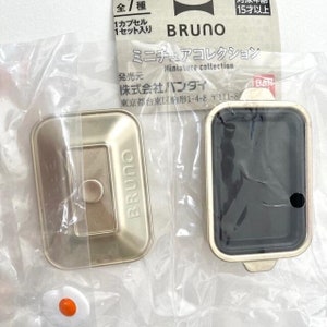 May include: A miniature gold-colored rectangular cooking appliance with a matching lid, featuring the brand name BRUNO embossed on the surface. The set includes a small white and orange fried egg accessory, all packaged in a clear plastic bag.