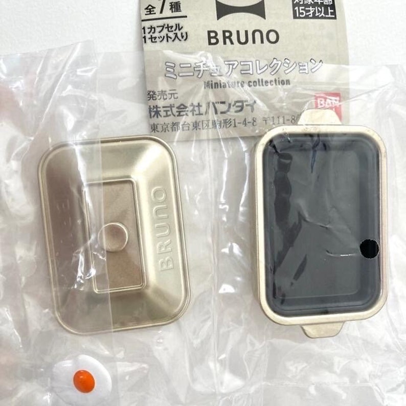 May include: A miniature gold-colored rectangular cooking appliance with a matching lid, featuring the brand name BRUNO embossed on the surface. The set includes a small white and orange fried egg accessory, all packaged in a clear plastic bag.