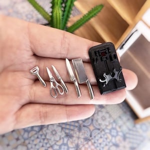 May include: A set of miniature kitchen knives and a pair of scissors, all made of silver metal. The knives and scissors are arranged on a black magnetic knife holder.