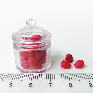 Może przedstawiać: Przezroczysty szklany słoik z pokrywką, wypełniony małymi czerwonymi malinami. Słoik znajduje się obok linijki pokazującej pomiary w centymetrach. Widocznych jest również kilka luźnych malin.