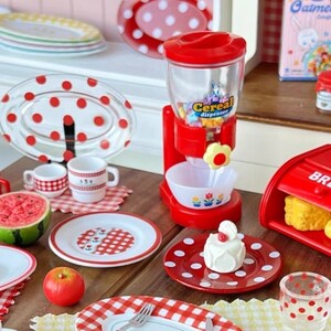 Op de afbeelding: Een rode en witte, met stippen versierd miniatuurkeuken speelset met een ontbijtgranen dispenser, borden, bekers en een kleine cake. De ontbijtgranen dispenser heeft een doorzichtige container met kleurrijke ontbijtgranen erin en een rode basis met de tekst "Ontbijtgranen dispenser" erop.