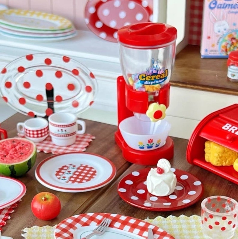 Op de afbeelding: Een rode en witte, met stippen versierd miniatuurkeuken speelset met een ontbijtgranen dispenser, borden, bekers en een kleine cake. De ontbijtgranen dispenser heeft een doorzichtige container met kleurrijke ontbijtgranen erin en een rode basis met de tekst "Ontbijtgranen dispenser" erop.