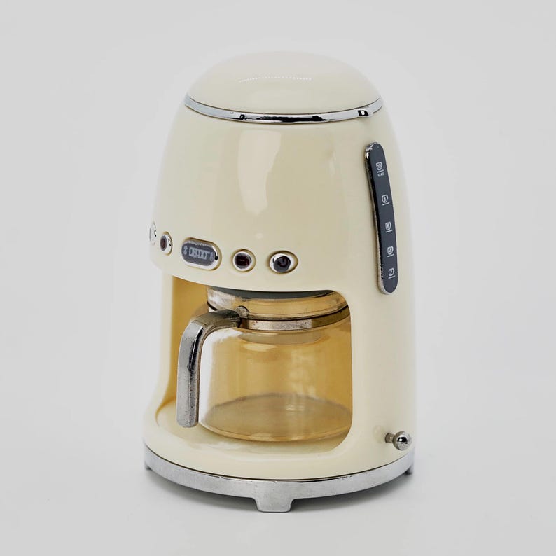 Peut inclure: Cafeti&egrave;re de style r&eacute;tro cr&egrave;me avec une carafe en verre. L'appareil dispose d'un affichage num&eacute;rique indiquant "08:00" et d'un indicateur de niveau d'eau. La cafeti&egrave;re a une garniture et une base chrom&eacute;es.