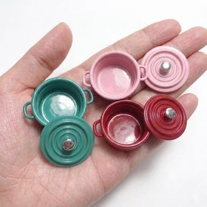 Könnte beinhalten: Miniatur-Kochgeschirr-Set in verschiedenen Farben. Das Set besteht aus teal-, rosa- und rotfarbenen Töpfen, jeweils mit passendem Deckel. Die Töpfe haben kleine Griffe und silberfarbene Knöpfe auf den Deckeln. Ideal für eine Miniaturküche.