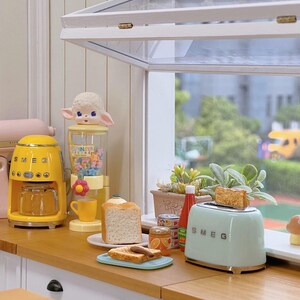 Peut inclure: Une sc&egrave;ne de cuisine miniature avec une cafeti&egrave;re jaune, un grille-pain vert menthe avec du pain grill&eacute; et un distributeur de bonbons surmont&eacute; d'une figurine d'agneau. Une tranche de pain et du pain grill&eacute; sont sur une assiette.