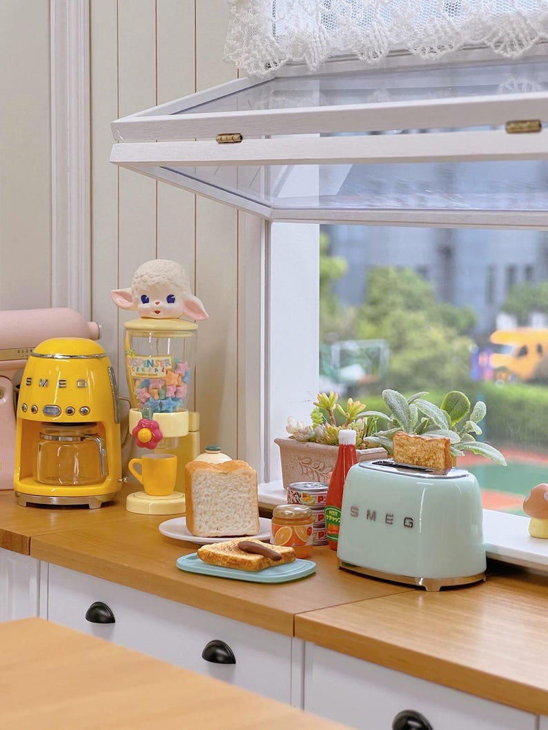 Peut inclure: Une sc&egrave;ne de cuisine miniature avec une cafeti&egrave;re jaune, un grille-pain vert menthe avec du pain grill&eacute; et un distributeur de bonbons surmont&eacute; d'une figurine d'agneau. Une tranche de pain et du pain grill&eacute; sont sur une assiette.
