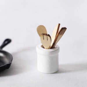 Puede incluir: Un porta utensilios de cerámica blanca con cuatro utensilios de cocina de madera en su interior. Los utensilios incluyen una cuchara, una espátula y dos tenedores.