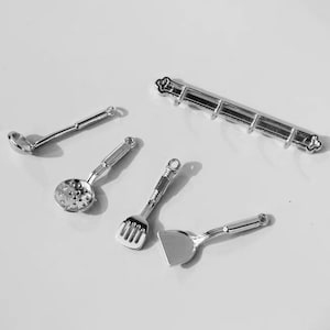 May include: A set of five miniature silver kitchen utensils, including a ladle, a slotted spoon, a spatula, and a turner, hanging on a silver rack.