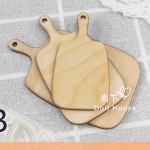 Puede incluir: Tres tablas de cortar de madera de diferentes tamaños, apiladas una encima de la otra. Las tablas son de color marrón claro y tienen una superficie lisa ligeramente texturizada. Las tablas están dispuestas en forma escalonada, con la tabla más grande en la parte inferior y la más pequeña en la parte superior. Las tablas están sobre una superficie a cuadros blancos y grises.