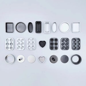 Może przedstawiać: Asortyment metalowych foremek i form do pieczenia w różnych kształtach, w tym okrągłych, kwadratowych, owalnych i w kształcie serca. Formy są ułożone na białej powierzchni.