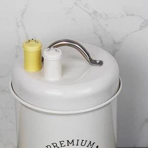 以下が含まれることがあります： シルバーの取っ手と「PREMIUM」という黒い文字が書かれた白い金属製の容器。容器には、黄色と白の蓋に小さな取っ手が付いています。