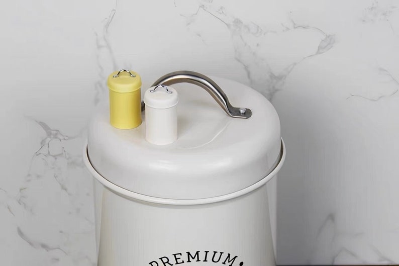以下が含まれることがあります： シルバーの取っ手と「PREMIUM」という黒い文字が書かれた白い金属製の容器。容器には、黄色と白の蓋に小さな取っ手が付いています。