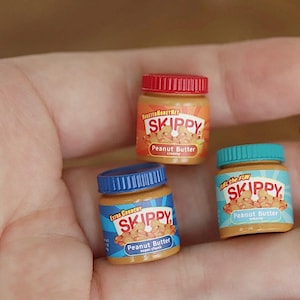 以下が含まれることがあります： スキッピーピーナッツバターのミニチュアジャーが3つ。ジャーには赤、青、ターコイズ色の蓋が付いています。各ジャーにはスキッピーのロゴと「ピーナッツバター」の文字が入ったラベルが付いています。ジャーには「クリーミー」、「エクストラクランチー」、「クリーミー」と表示されています。