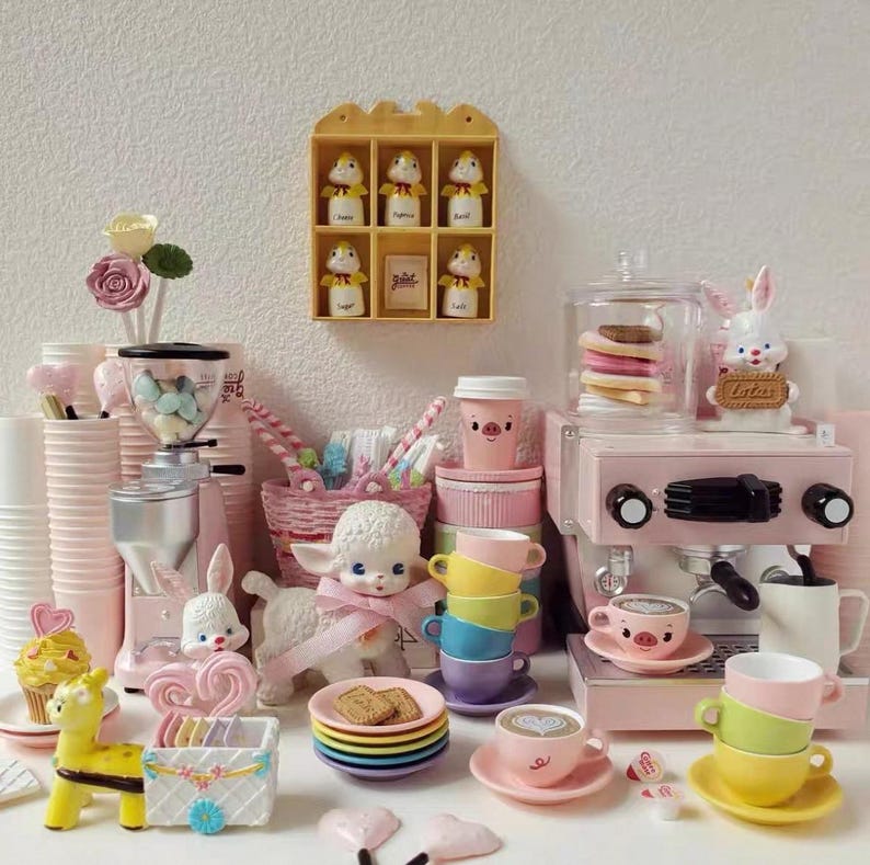 Pode incluir: Uma cole&ccedil;&atilde;o de miniaturas e acess&oacute;rios cor-de-rosa e pastel com tema de cozinha e caf&eacute;. Inclui uma m&aacute;quina de caf&eacute; expresso de brinquedo, x&iacute;caras, pires, um moedor de caf&eacute; e figuras decorativas de animais e sobremesas.