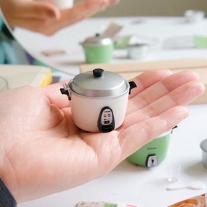 May include: A small white rice cooker with a silver lid and black buttons. The cooker is being held in a hand.