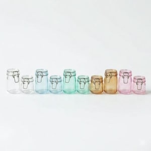 Può includere: Una collezione di otto barattoli di vetro con chiusure metalliche in varie dimensioni e colori, tra cui trasparente, blu, verde e ambra. I barattoli sono disposti in fila su sfondo bianco.