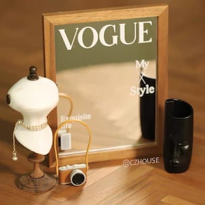 May include: A wooden framed mirror with a Vogue magazine reflection. The reflection shows the text "My Style" and the text "Exquisite Life". The mirror is sitting on a wooden surface with a small white mannequin, a miniature camera, and a black vase.