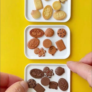 Könnte beinhalten: Eine Sammlung von Miniaturkeksen und -gebäck, angeordnet auf weißen Tabletts vor einem gelben Hintergrund. Die Kekse umfassen verschiedene Formen wie Teddybären, Muscheln, Lebkuchenmänner und runde Kekse. Das Bild ist gut beleuchtet und zeigt die Details jedes Kekses.