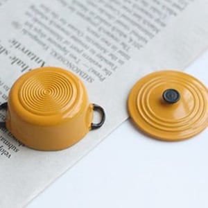 May include: A miniature mustard-yellow cooking pot with black handles and a matching lid. The pot has a circular design on the bottom and the lid has a black knob. The items are on a newspaper background.