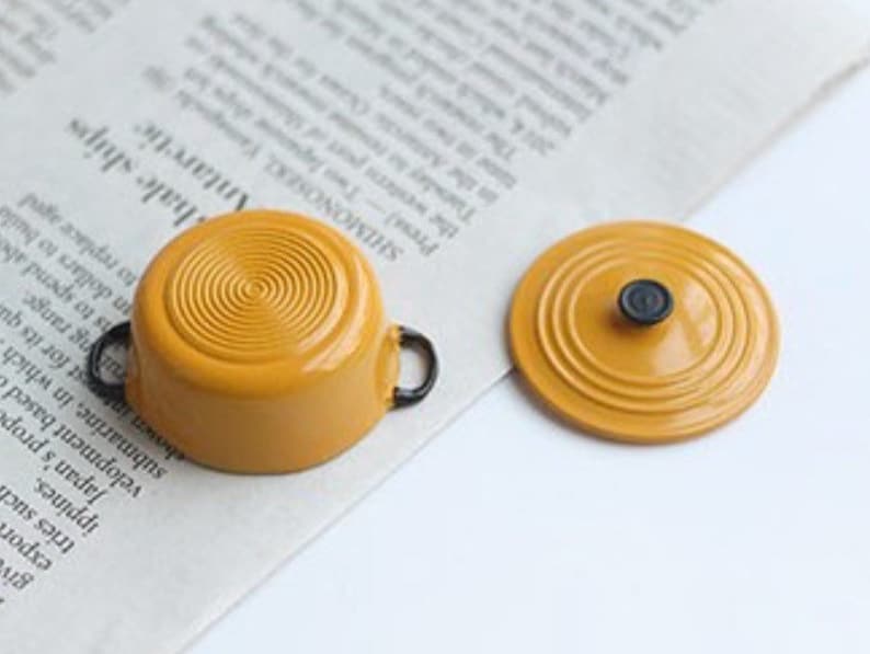 May include: A miniature mustard-yellow cooking pot with black handles and a matching lid. The pot has a circular design on the bottom and the lid has a black knob. The items are on a newspaper background.